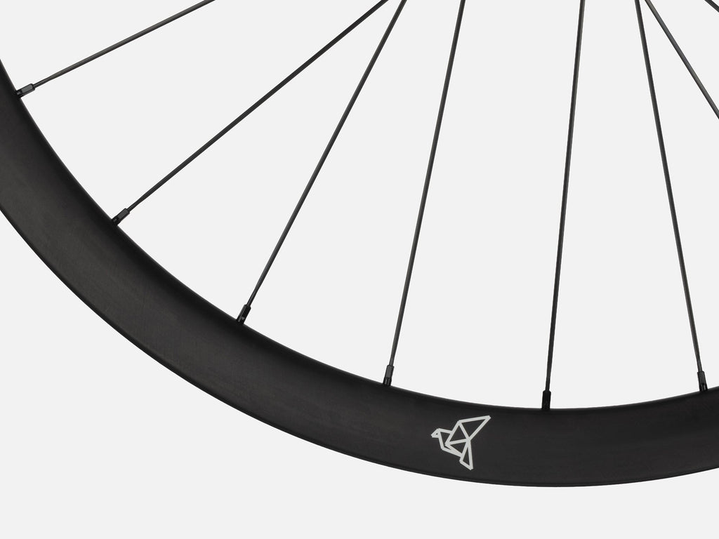 Close-up of Litebike Arc-35 carbon wheelset. A black bicycle wheel rim with thin black spokes and a small white geometric logo near the top, shown against a light gray background.